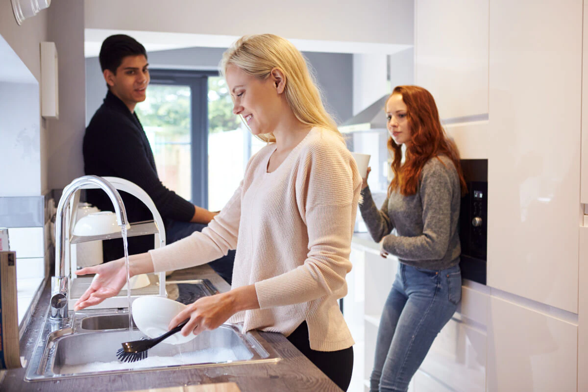 Woman washing the dishes while her roommates watch her.