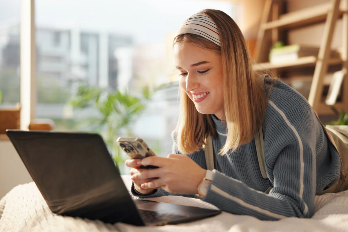 Woman laying down on her bed texting on her phone next to her laptop.