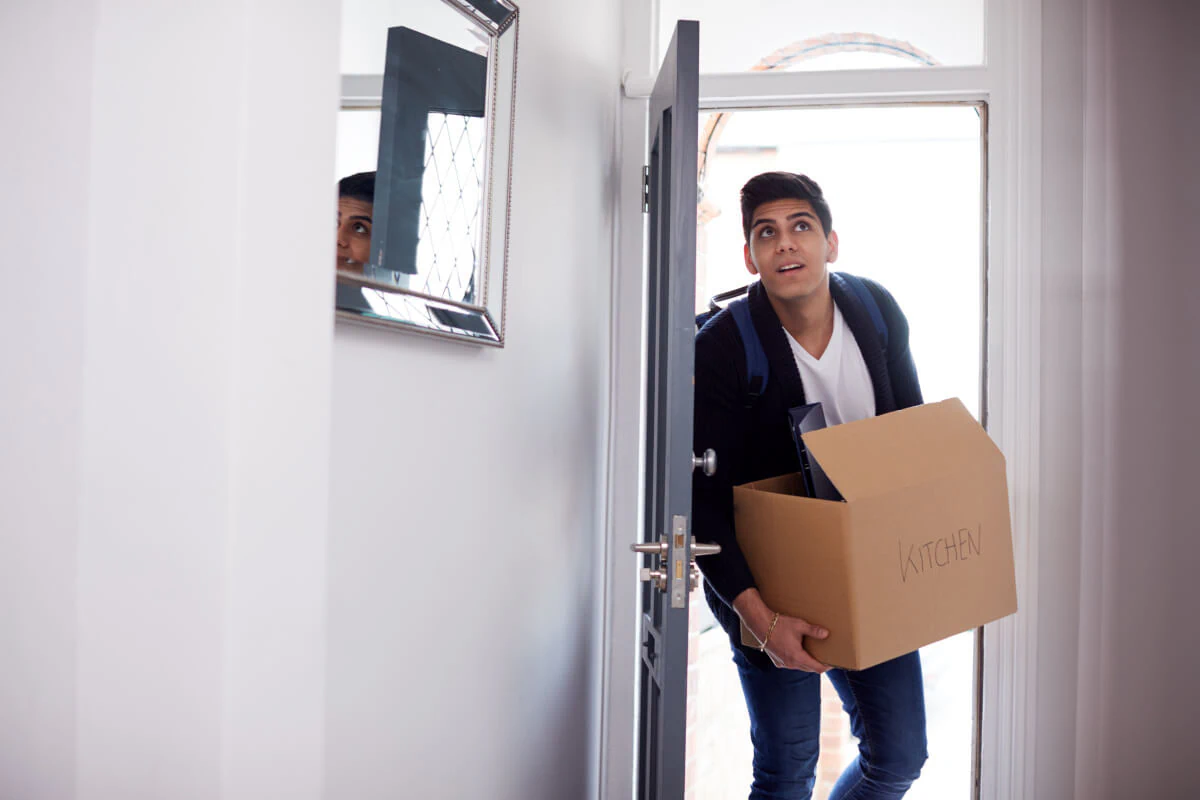 College student entering his new apartment and carrying a box labelled Kitchen.